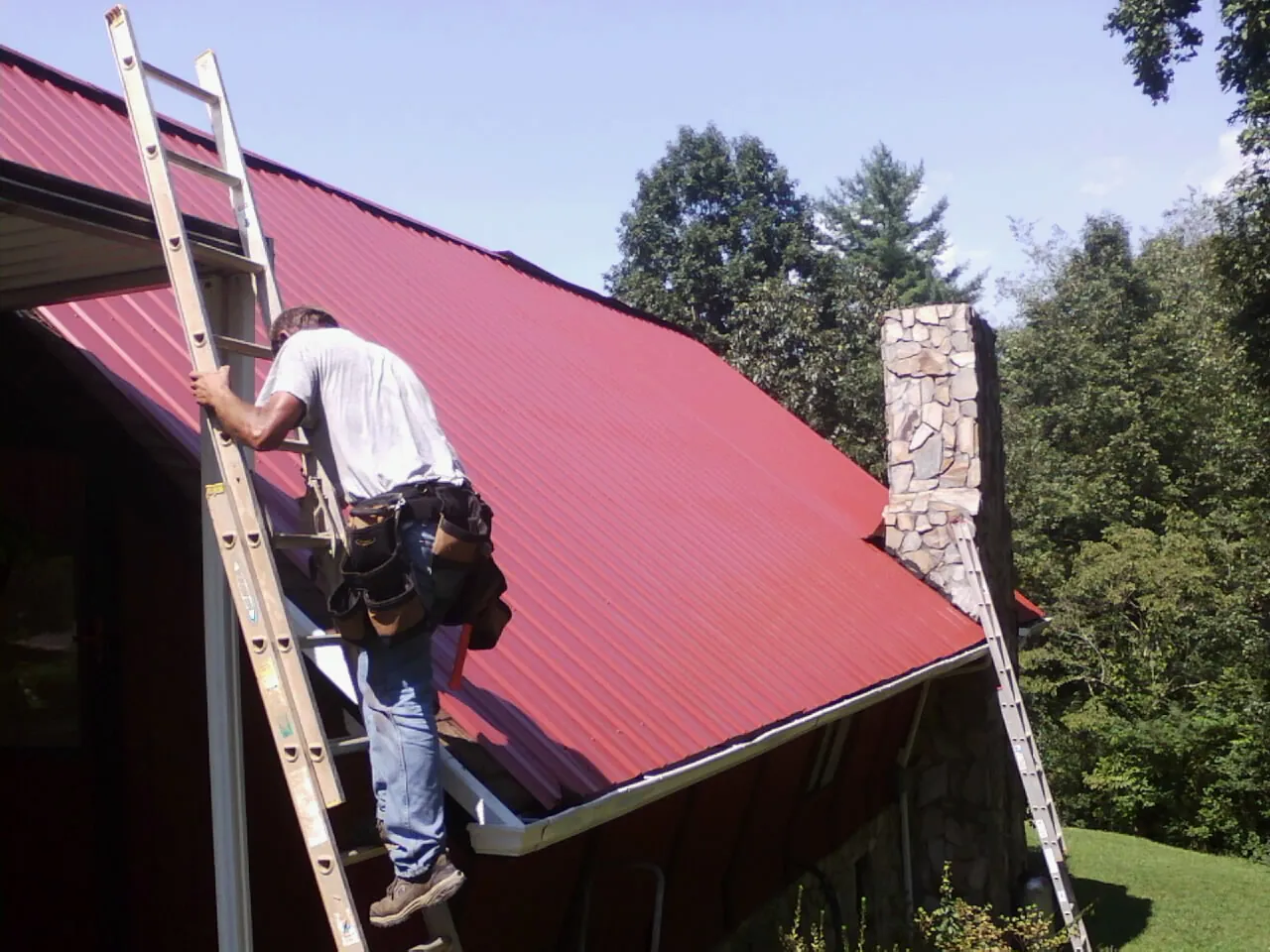 Roofer on a ladder beside a red metal roof and stone chimney — Soffit & Fascia Repair by Roofing Brothers in East Aurora